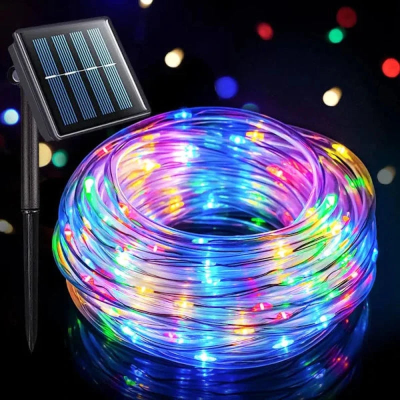 A coiled length of flexible rope light with multiple colored LED bulbs is shown with its attached solar panel and ground stake against a dark background with colorful bokeh lights. The rope light displays a mix of blue, pink, yellow, and white lights.