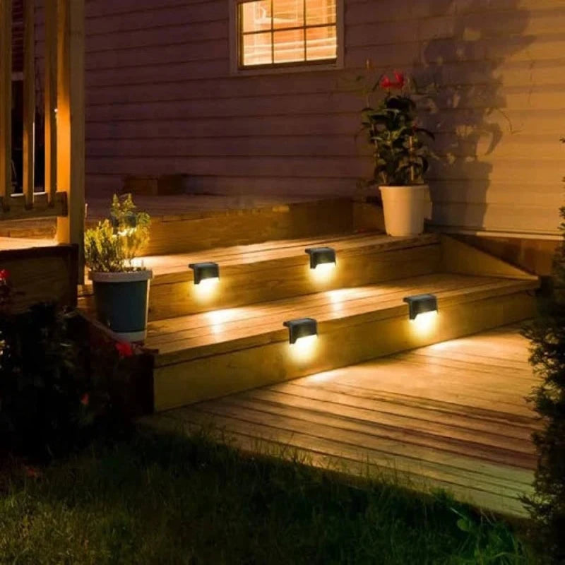 The image shows an outdoor staircase at night, illuminated by four small, dark, rectangular solar lights mounted on the sides of the steps. The lights cast a warm white glow downwards, lighting up the steps. The steps are made of wood and lead up to a house. A window of the house is lit, and there are plants in pots nearby. Grass is visible at the bottom of the steps.