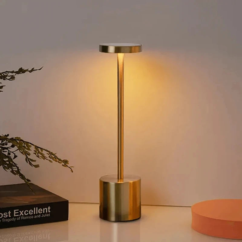 A cordless, rechargeable LED table lamp with a brushed gold finish, featuring a cylindrical base, a slender stem, and a circular light panel emitting a warm glow, positioned on a light-colored surface next to a book and a round orange object.