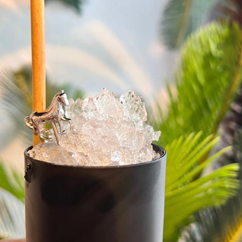 A cocktail in a black cup filled with crushed ice and a silver horse-shaped cocktail pick, with a blurred tropical background.