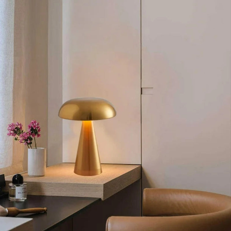 A gold, mushroom-shaped lamp illuminates a light wooden desk in a warm-toned room, casting a soft glow on a small vase of pink flowers beside it, with a brown leather armchair partially visible in the foreground, suggesting a cozy and inviting atmosphere.