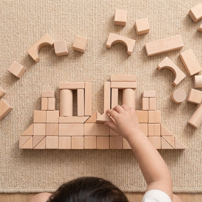 Overhead view of a child's hand placing a triangle block into a temple-like structure with columns and arches, on a woven rug