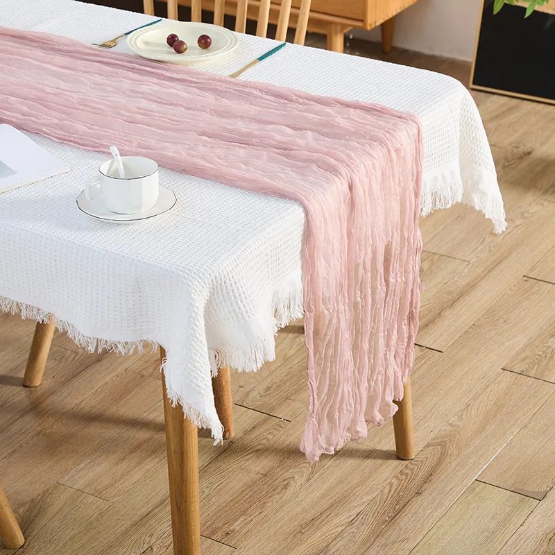 A blush pink gauze table runner with a soft, crinkled texture draped over a white textured tablecloth on a wooden dining table, set with a teacup and saucer, a plate with grapes, and gold-accented cutlery.