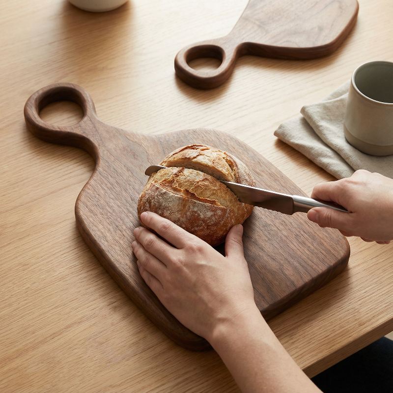 Person slicing bread on a wooden cutting board with a knife, on a wooden table.