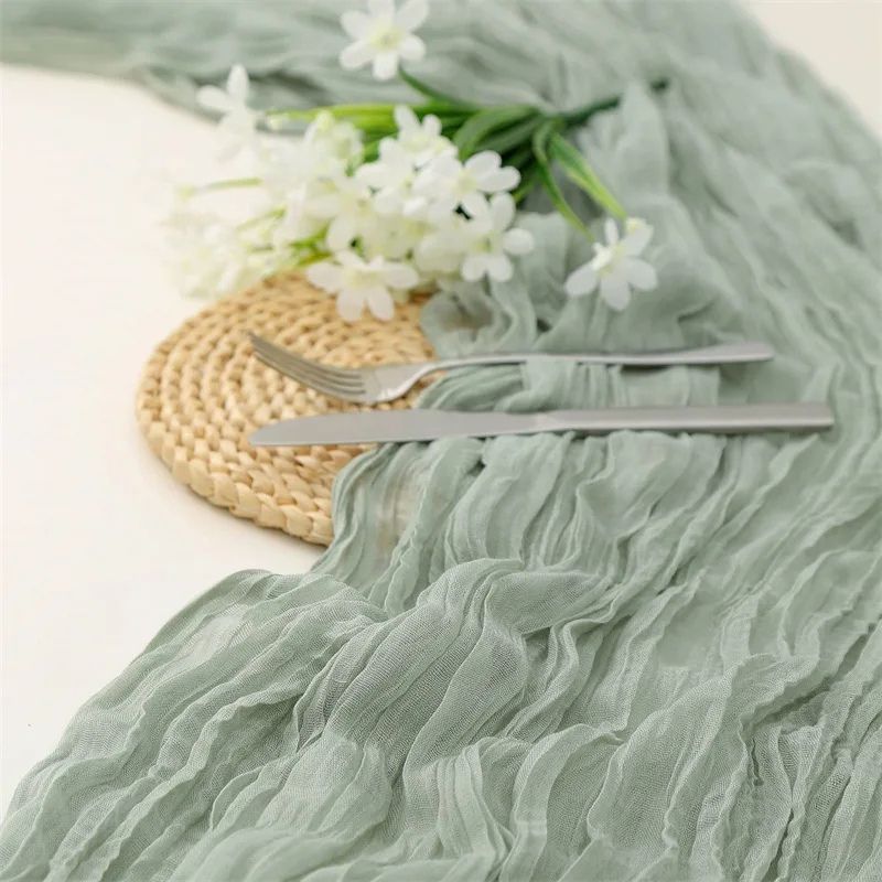 A sage green gauze table runner with a crinkled texture elegantly draped on a white table, accompanied by a woven placemat, silverware, and delicate white flowers.