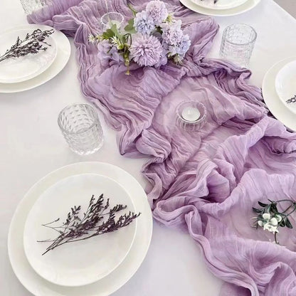 A soft lilac gauze table runner with a crinkled texture elegantly draped across a white table, adorned with pastel purple flowers, crystal glassware, white plates, and delicate dried floral accents.