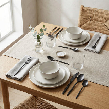 Dining table set with white ceramic dishes, black cutlery, and glasses on a wooden table.