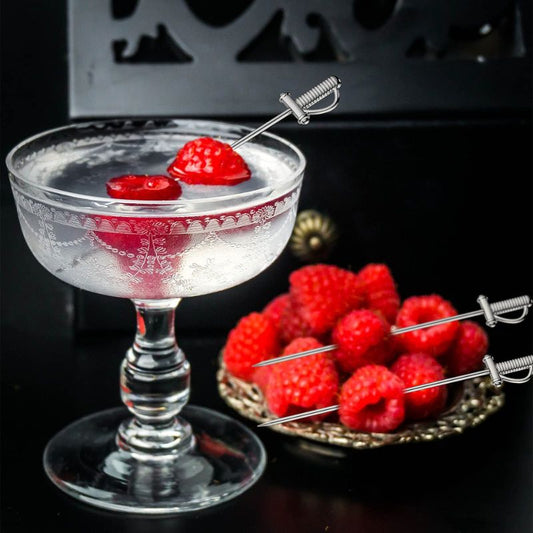 A clear cocktail in a vintage coupe glass garnished with two raspberries on sword-shaped stainless steel cocktail picks. A small dish of raspberries and two more cocktail picks are visible in the background.
