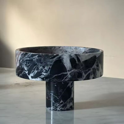 A black and white marbled pedestal bowl sits on a light gray marble surface, offering a modern twist with its high-footed design. The softly lit background highlights the bowls smooth, polished texture and intricate marbling patterns, making it an exquisite premium fruit bowl.