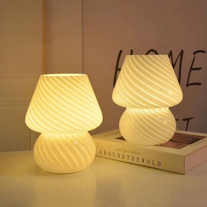 Two dimmable mushroom lamps with a striped, translucent Murano-style glass design emit a warm, ambient glow. Placed on a white surface, one lamp rests atop a hardcover book, adding a decorative touch. The soft lighting enhances the cozy and modern aesthetic of the space.