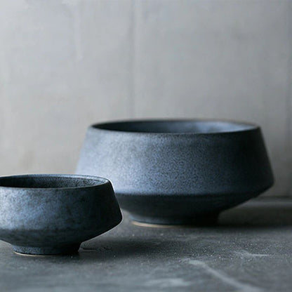 Two minimalist ceramic bowls with a matte, dark gray finish on a textured surface against a neutral-toned background. The smaller bowl is in the foreground, while the larger one is slightly behind.