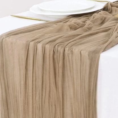 A soft beige gauze table runner with a delicate, crinkled texture elegantly draped over a white table, complemented by stacked white plates and gold-accented flatware.