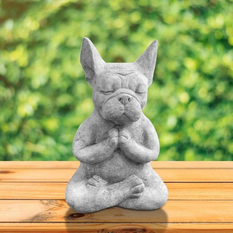 A gray, stone-like statue of a French Bulldog meditating in a lotus position sits on a wooden surface against a blurred green background. The bulldog has its eyes closed and paws pressed together in a prayer-like gesture.