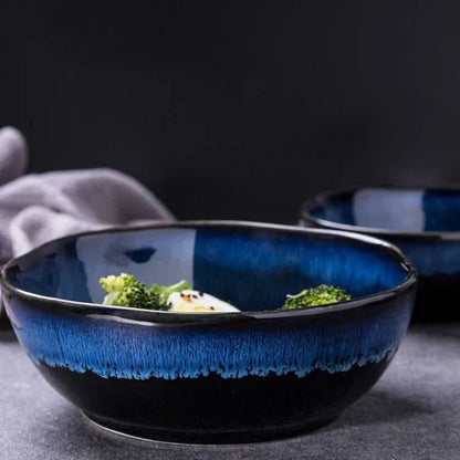 Black and blue ceramic wave bowl with a textured glaze, partially filled with a fresh salad, set against a dark background with another matching bowl in the distance