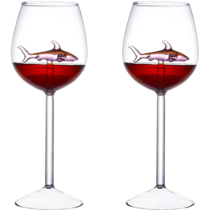 A pair of wine glasses filled with red liquid, each containing a small, pink glass shark figurine.