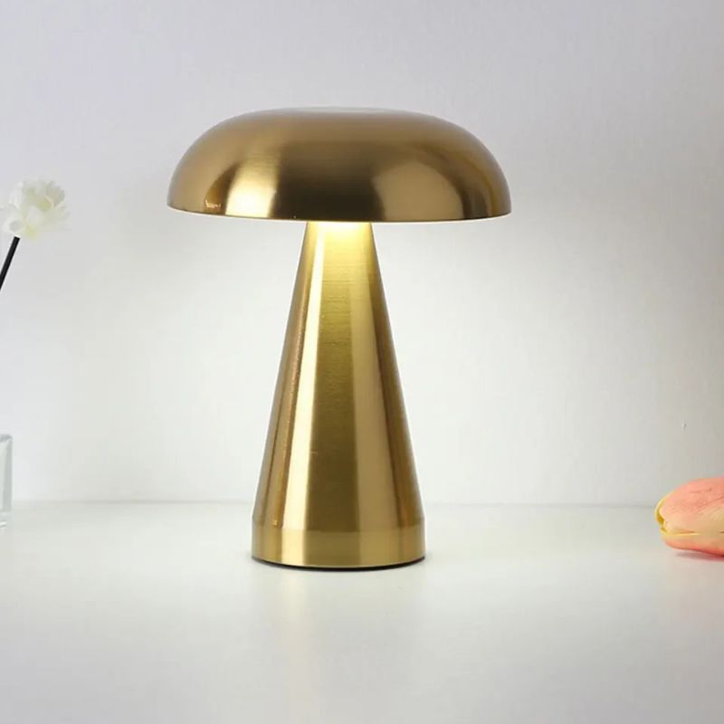 A gold, mushroom-shaped lamp with a glowing light beneath its dome sits on a white surface, with a blurry white flower to the left and a partially visible peach-colored object to the right, all set against a soft, light background.