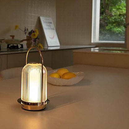 A lit, portable, lantern-style lamp with a vertical-ribbed glass shade and a gold-toned base, sitting on a kitchen counter next to a bowl of lemons.