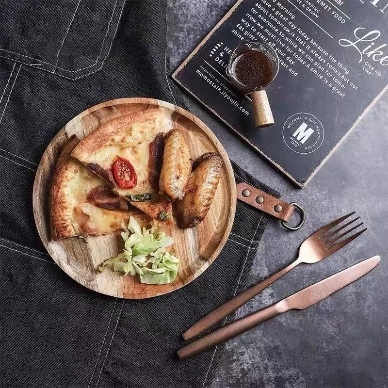 A small round wooden plate holds a slice of pizza, a few pieces of chicken wings, and a small side of salad. A set of rose gold cutlery and a menu are also visible on the dark tablecloth.