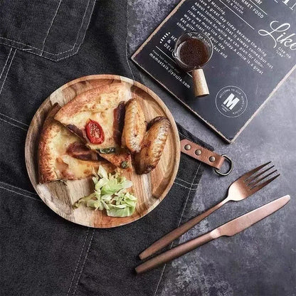 A small round wooden plate holds a slice of pizza, a few pieces of chicken wings, and a small side of salad. A set of rose gold cutlery and a menu are also visible on the dark tablecloth.