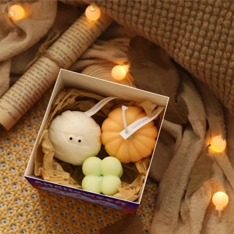 A gift box contains three Halloween-themed candles nestled in brown crinkle-cut paper. Inside are a white ghost-shaped candle with two black dot eyes, an orange pumpkin-shaped candle, and a light green four-leaf clover-shaped candle. Each candle has a white ribbon tag. The box sits on a textured surface with soft, warm-toned string lights scattered around it. A rolled-up piece of paper with printed text is visible in the upper left corner.