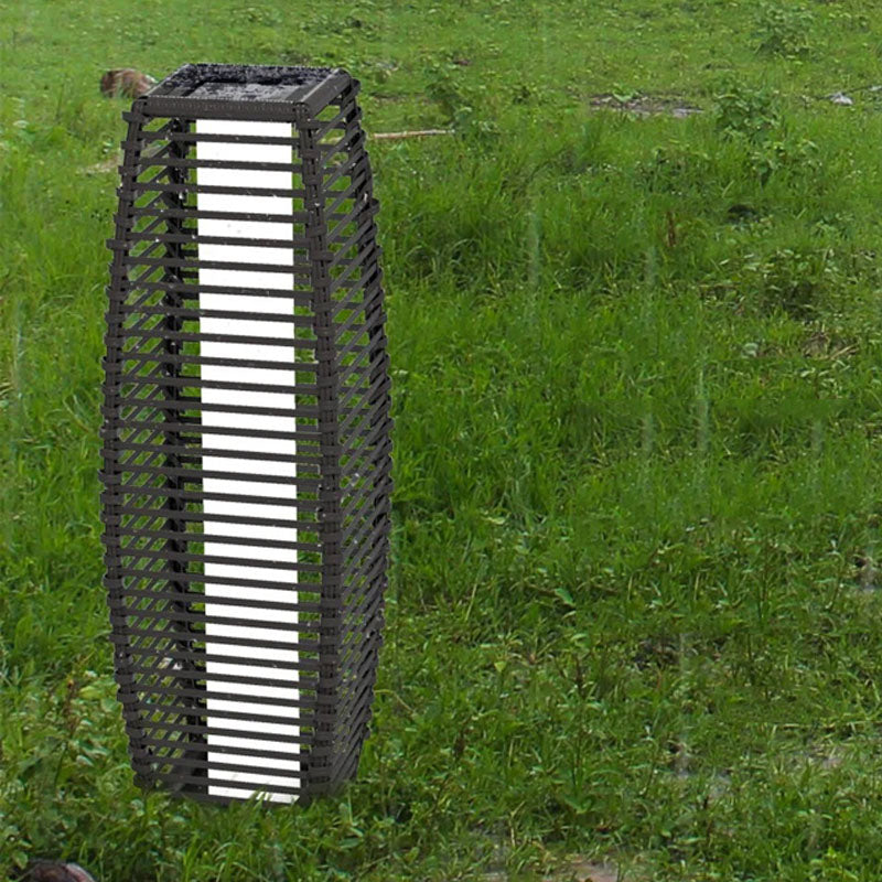 A tall, cylindrical outdoor lantern with a slatted black frame stands in a grassy field in the rain.