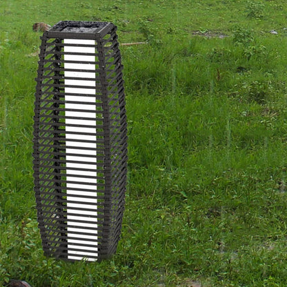 A tall, cylindrical outdoor lantern with a slatted black frame stands in a grassy field in the rain.