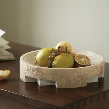 A travertine stone bowl with a geometric design cradles green pears, a yellow apple, and a halved citrus fruit. Nestled on a dark wooden table against a light green wall, the scene exudes organic texture. Some papers are partially visible on the left, adding an element of home styling.