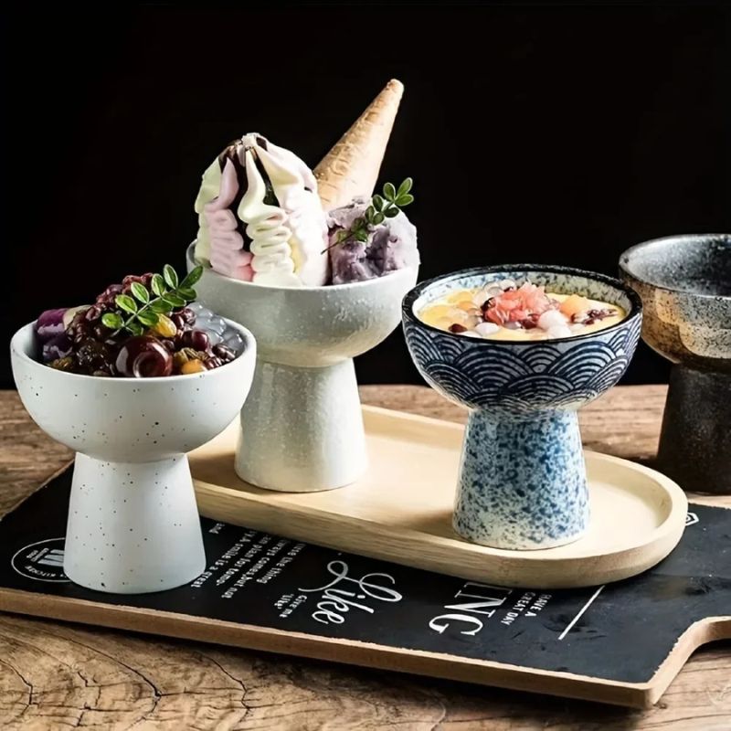 A set of ceramic pedestal bowls in various colors and textures, elegantly arranged on a wooden tray. Each bowl contains a dessert, including ice cream, sweet beans, and fruit toppings. The background features a dark, moody setting with a wooden surface.