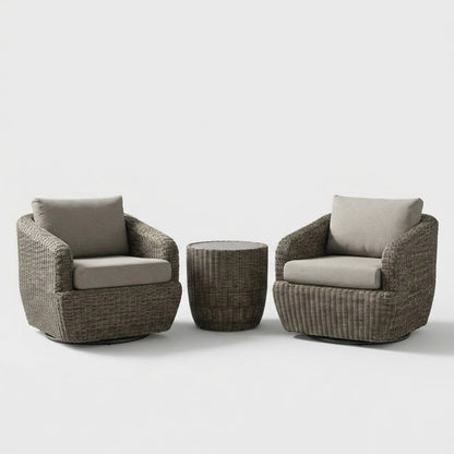 Two gray-brown rattan swivel barrel chairs with greige cushions and dark round swivel bases flanking a round rattan drum table with a pale gray glass top on a plain studio backdrop
