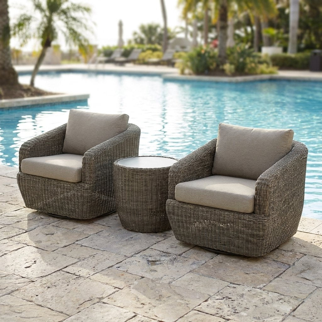 Two gray-brown rattan swivel barrel chairs with greige cushions and a round rattan table on a resort pool deck with a turquoise pool and palm trees
