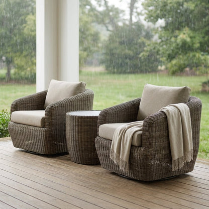 Two gray-brown rattan swivel barrel chairs with greige cushions and a round rattan table on a covered wooden porch in the rain, with a fringed cream throw over the right chair