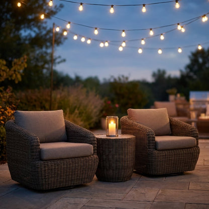 Two gray-brown rattan swivel barrel chairs with greige cushions and a round rattan table with a lit white pillar candle in a glass hurricane on a stone patio at dusk under string lights