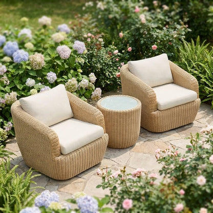 Raised view of honey-tan rattan swivel barrel chairs with cream cushions and a round rattan table on flagstones among hydrangeas, roses and ferns