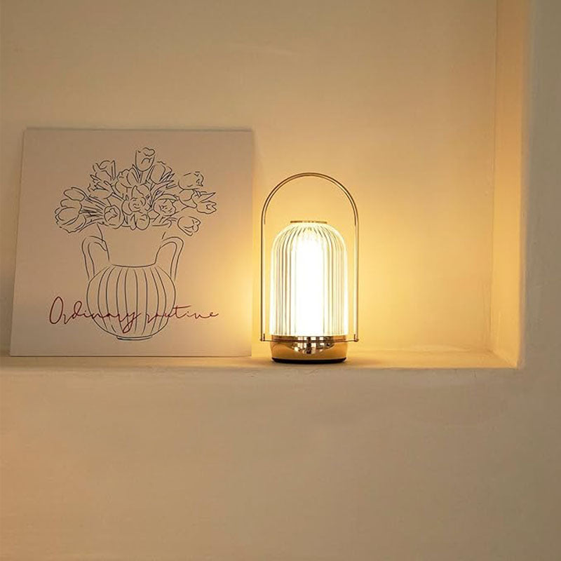 A glowing modern lantern lamp with a vertical-ribbed glass shade and a gold base, placed on a white shelf next to a floral line-art print.