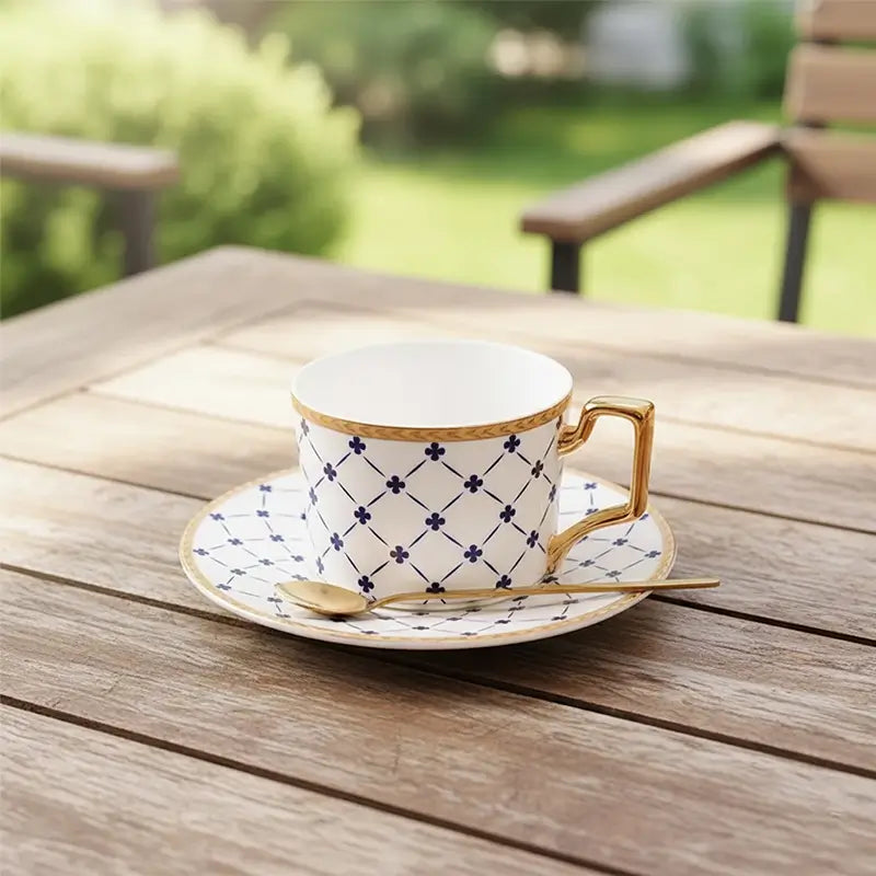 White teacup and saucer with blue lattice and clover pattern, gold trim, and a gold spoon on an outdoor wooden table.