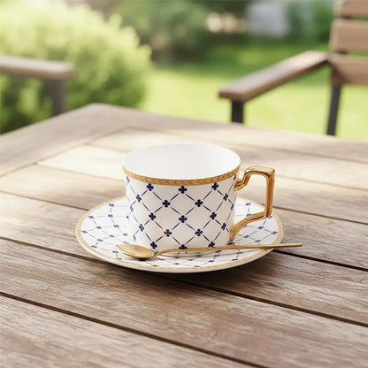 White teacup and saucer with blue lattice and clover pattern, gold trim, and a gold spoon on an outdoor wooden table.