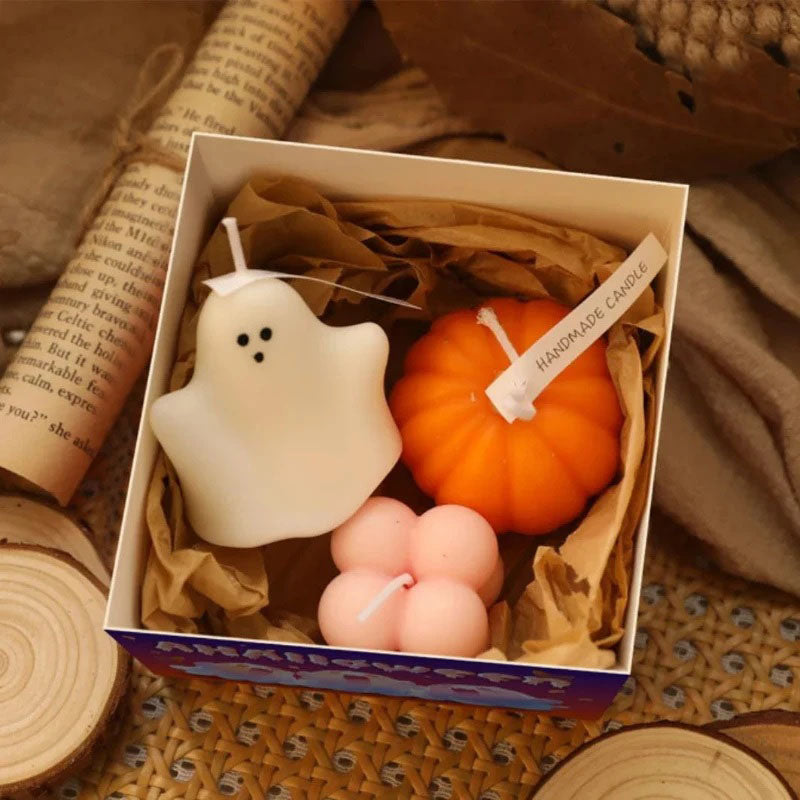 A gift box contains three Halloween-themed candles nestled in brown crinkle-cut paper. Inside are a white ghost-shaped candle with two black dot eyes, an orange pumpkin-shaped candle with a white string tied around it and a "Handmade Candle" tag, and a light pink four-leaf clover-shaped candle. The box sits on a textured surface with soft, warm-toned string lights scattered around it and wooden slices nearby. A rolled-up piece of paper with printed text is visible in the upper left corner.