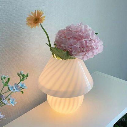 A glowing striped glass mushroom lamp doubles as a vase, holding a pink hydrangea and a yellow gerbera daisy. The soft, ambient lighting enhances the delicate floral arrangement, creating a cozy and artistic decor piece on a white surface against a neutral-toned wall.