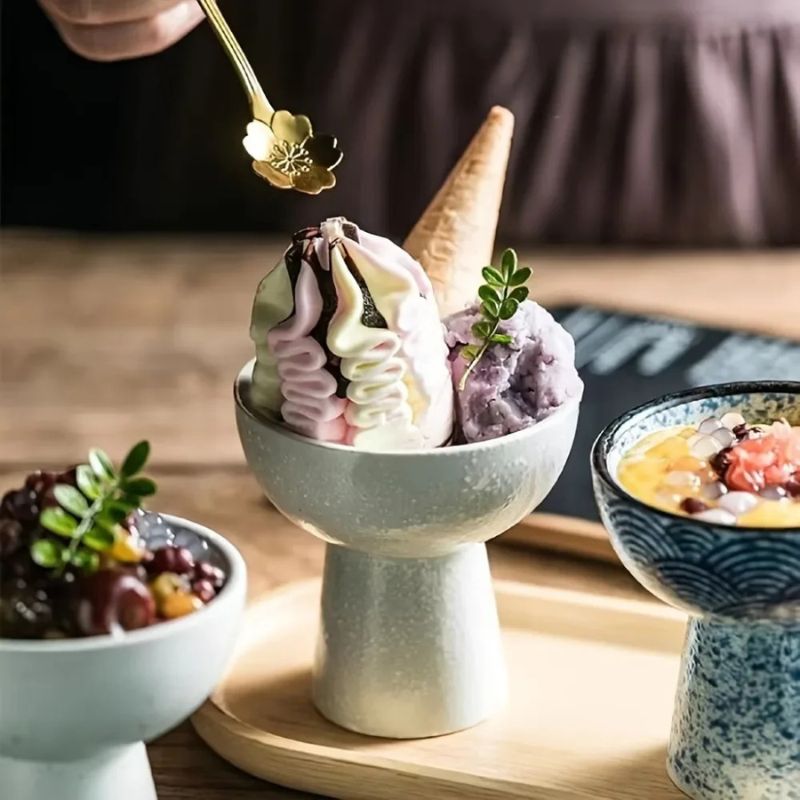 Elegant dessert in a ceramic pedestal bowl, featuring pastel swirled soft-serve with chocolate drizzle, a waffle cone, and a scoop of taro. Garnished with greenery, with other colorful desserts in the background.