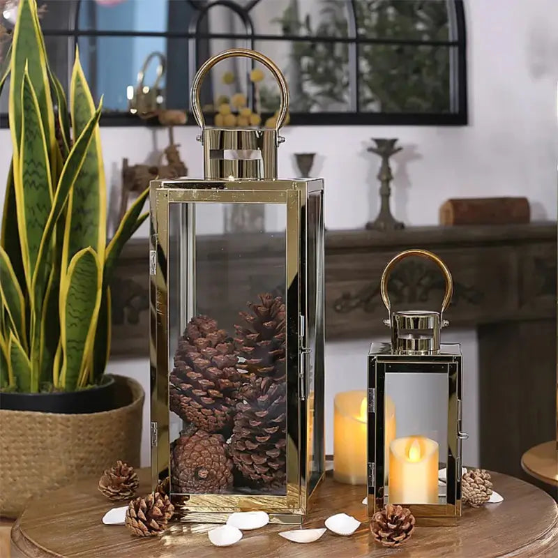 Two rectangular gold-toned stainless steel and glass lanterns used for decoration with pinecones and LED candles.
