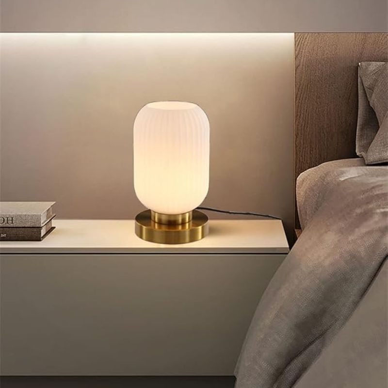 A lit, ribbed white glass table lamp with a gold base sits on a white nightstand next to a stack of books and a bed with neutral-toned bedding. A warm light strip illuminates the wall behind the nightstand. This could be an Auwieou Mid Century Modern Table Lamp.