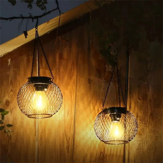 Two black metal mesh solar lanterns hanging from tree branches, glowing warmly against a wooden wall at night. The lanterns emit a soft, ambient light, creating a cozy and decorative outdoor atmosphere. Their intricate mesh design allows the light to cast beautiful patterns, enhancing the aesthetic appeal of the space.