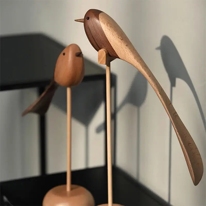 Two decorative wooden bird figurines with long tails on wooden stands; the front bird is two-toned, and the rear bird is a lighter wood.