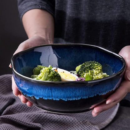 Person holding a black and blue ceramic wave bowl filled with a fresh salad, featuring broccoli, lettuce, and a sliced boiled egg. The glossy finish and textured glaze of the bowl stand out against a dark background.