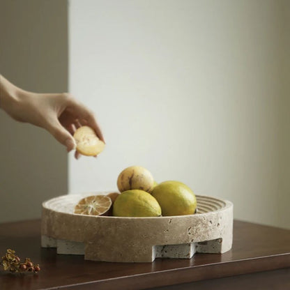 A hand places a lemon wedge on a low, round travertine stone bowl filled with whole lemons. The bowl rests on a wooden surface against a plain wall, complemented by the organic texture of a nearby plant piece. The soft lighting enhances the serene home styling atmosphere.