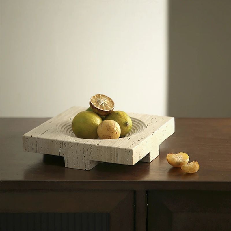 A square travertine stone tray with concentric circles holds several limes and a halved lime on top, placed on a wooden surface. Two lime halves rest beside the tray, highlighting its natural beauty. The scene is softly lit, offering an elegant touch to home styling.