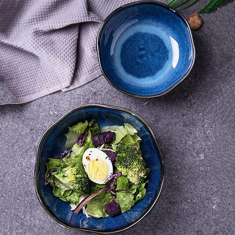 Two blue ceramic wave bowls with a glossy finish, one filled with a fresh salad topped with a boiled egg, placed on a textured grey surface with a napkin nearby.