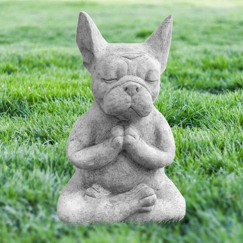 A gray, textured statue of a French Bulldog in a meditative yoga pose sits on green grass. The dog has its eyes closed and its front paws pressed together in front of its chest.