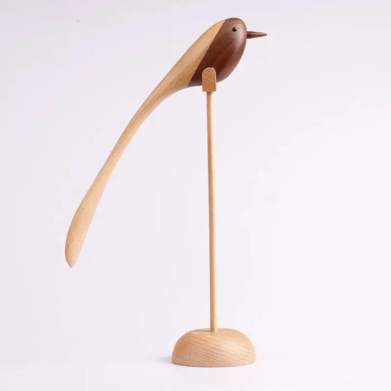 A single decorative wooden bird figurine with a long, sweeping tail, made from contrasting light and dark wood, standing on a thin wooden dowel and round base against a white background.
