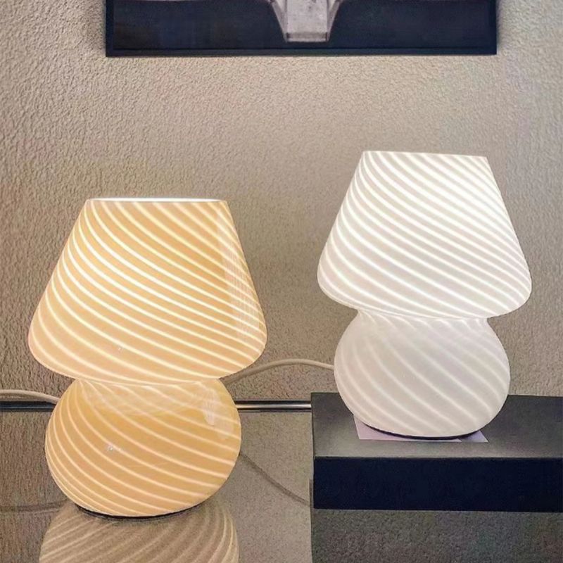 Two striped glass mushroom lamps emit a warm and cool glow, placed on a reflective glass surface against a textured wall. The soft lighting and modern design create a stylish and cozy ambiance, perfect for bedside or accent lighting.